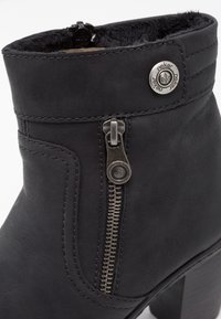 Bota de tobillo de cuero negro con cremallera lateral, hardware decorativo de plata y un suave forro de piel en el cuello. Detalles de costura destacados.