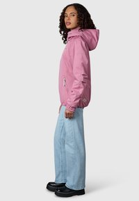 Vrouw met krullend haar, een roze gewatteerde jas met capuchon, lichtblauwe wijde jeans en zwarte plateauzolen, staand zijwaarts.