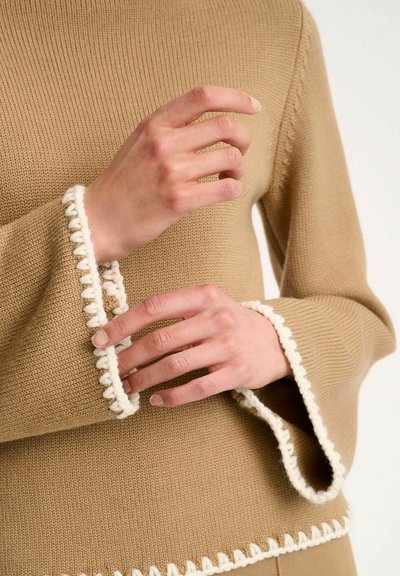 Pull en tricot beige avec des manches évasées et des bordures en dentelle blanche, mettant en avant un tissu texturé et une coupe décontractée.