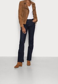 Tan suede biker jacket, white lacy top, dark blue flared jeans, and brown heeled ankle boots, standing against a neutral background.