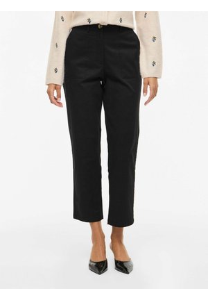 Kobieta ubrana w czarne, proste spodnie o długości 7/8, beżowy sweter z kwiatowym haftem oraz czarne szpilki w szpic, stojąca na białym tle.