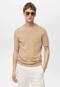 Pulover tricotat bej cu mâneci scurte și guler rotund, dintr-un material texturat. Modelul poartă ochelari de soare maro și pantaloni albi. Fundal neutru.
