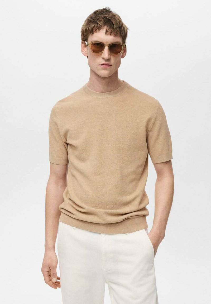Beige pletený svetr s krátkým rukávem a kulatým výstřihem, texturovaná tkanina. Model má na sobě hnědé sluneční brýle a bílé kalhoty. Neutrální pozadí.
