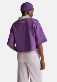 Camiseta morada recortada con cuello gris y logo de "Stranger Things" en rosa. Combinada con pantalones claros a rayas, mostrando un estilo casual.