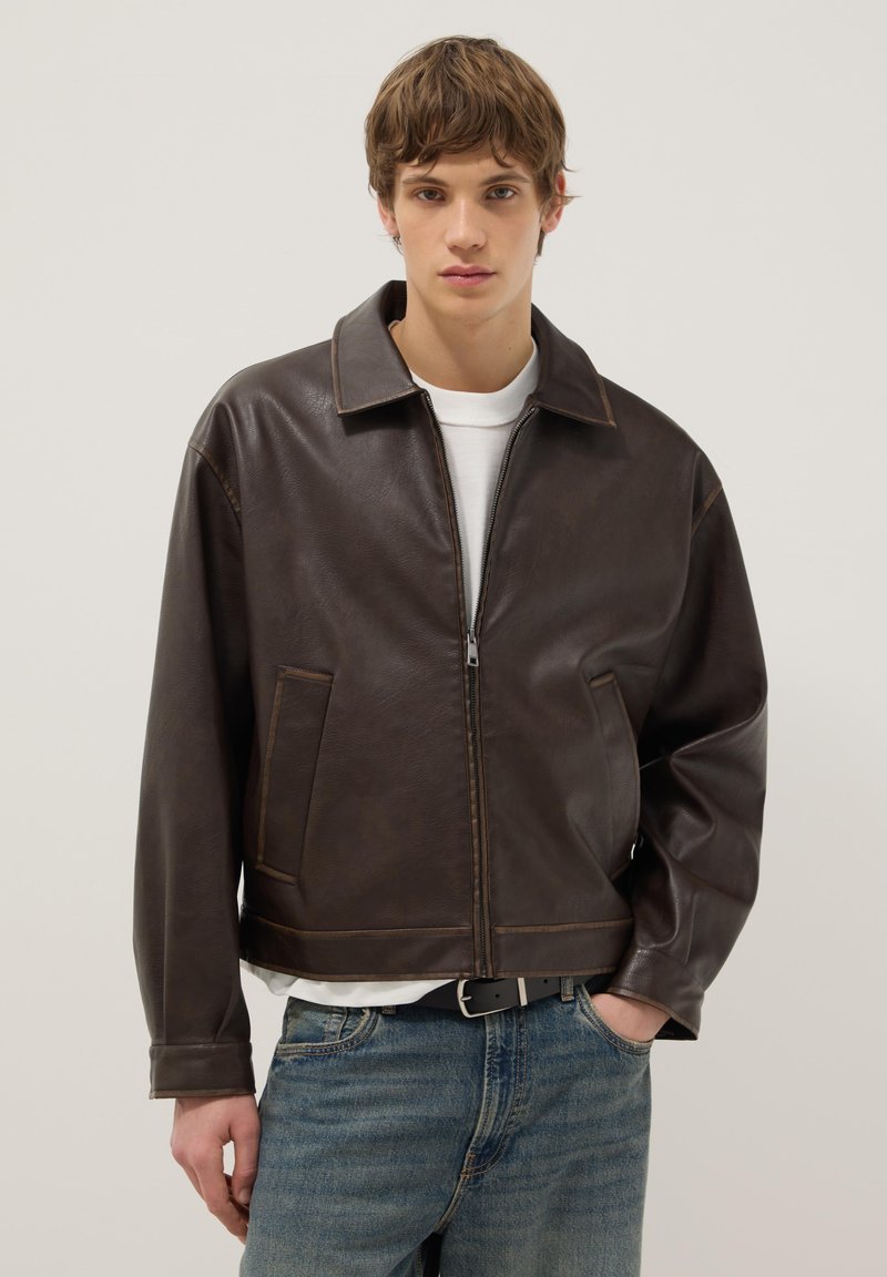 Young person wearing a dark brown leather zip-up jacket over a white shirt and blue jeans, standing with one hand in pocket.