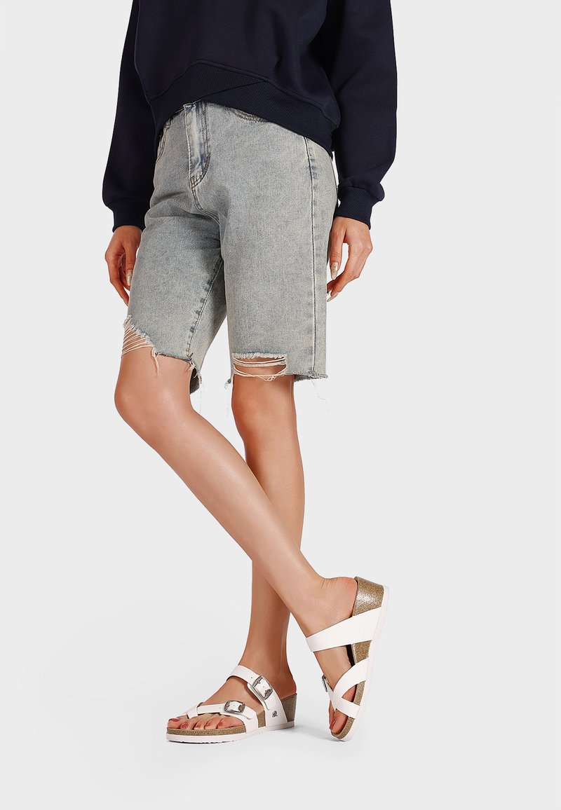 Sandali bianchi con accessori in argento e dettagli in oro, abbinati a pantaloncini di denim chiaro con orli sfrangiati. L'outfit ha un aspetto casual.