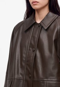 Femme portant une veste en cuir marron foncé avec un grand col et des boutons-pression recouverts, montrée des épaules jusqu'au milieu du torse.