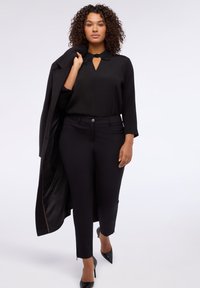 Blusa nera con colletto decorativo, pantaloni neri aderenti e décolleté neri. Modella tiene un cappotto nero, con uno sfondo chiaro.
