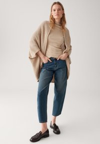 Cardigan en tricot beige superposé sur un col roulé côtelé beige clair, associé à un jean loose bleu moyen et des mocassins sombres. Textures lisses et coupe décontractée.