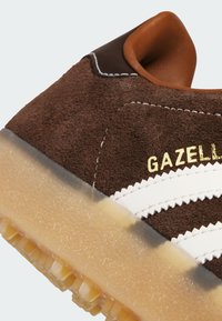 Baskets en daim brun avec des rayures en cuir blanc et des lettres dorées. Dotées d'une semelle en gomme semi-transparente avec un motif texturé.