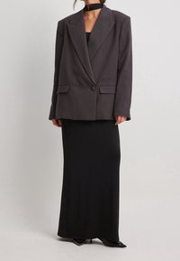 Mujer vistiendo un blazer oversized de color gris oscuro, top negro con choker, falda negra larga y zapatos de punta negros frente a un fondo sencillo.