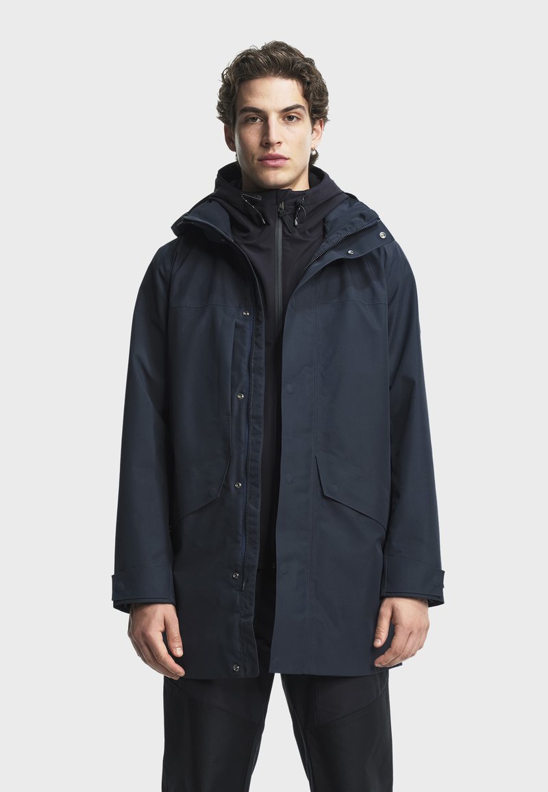 Marinblå vattentät jacka med huva, försedd med tryckknappar, fyra framfickor och en slät textur, lagd över en svart zip-up tröja.