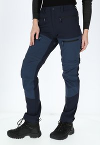 Technical outdoor pants in dark blue and navy fabric, featuring multiple zippered pockets and reinforced knee sections. Textured material.