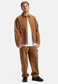 Braune Baumwollarbeitsjacke mit zwei Taschen, kombiniert mit passenden braunen Cargo-Hosen mit seitlicher Tasche. Getragen mit schwarzen Schuhen.
