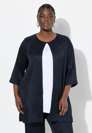 Veste texturée bleu marine avec un décolleté arrondi, des manches trois-quarts et une fermeture à un bouton, portée sur un haut blanc.