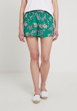 Vrouw met groene bloemenprint korte broek, wit shirt en witte instappers, staand op een lichte achtergrond.