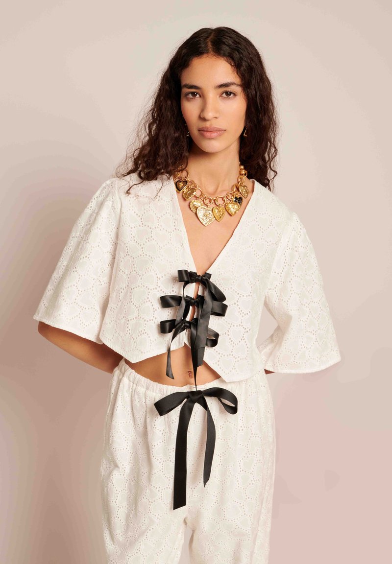Top cropped branco com mangas curtas, padrão de renda com ilhós, laços em fita preta no decote, combinado com calças a condizer. Colar dourado em forma de coração.