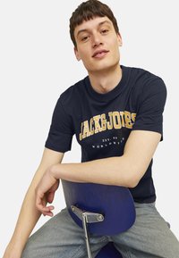 Navy blue cotton t-shirt with white and yellow "JACK&JONES" logo text, round neckline, short sleeves, worn with light blue jeans.