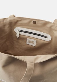 Beige tote-väska med en texturerad kanvasutsida. Inuti finns en vit dragkedjeficka och en logotagskylt. Översta stängningen är öppen.