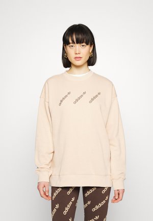 Lys pink sweatshirt med ribbet runde halsudskæring, oversized pasform og gentagne mørkebrune "adidas" logoer. Sammen med brune mønstrede leggings.