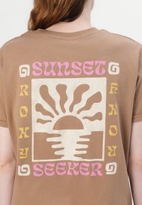 Brown cotton t-shirt featuring a graphic design with the text "SUNSET SEEKER" and a sun motif in beige, yellow, and pink colors.
