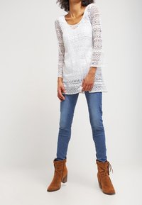 Top bianco in pizzo con maniche lunghe a campana e motivi geometrici. Abbinato a jeans skinny blu e stivali alla caviglia marroni con tacco spesso.