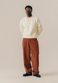 Mężczyzna w okularach, w kremowej bluzie z napisem "ILLINIS", rdzawych spodniach w kratę, czarnych butach i dwóch złotych naszyjnikach, stojący z rękami za plecami.