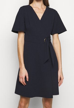 Femme portant une robe noire longueur genou avec des manches volantées, un décolleté en V et une ceinture nouée à la taille avec un détail d'anneau doré.