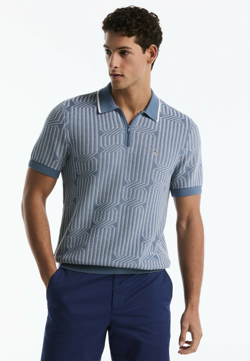 Man wearing a blue patterned short-sleeve polo shirt with a zip collar and navy blue pants, looking to his left with hands in pockets.
