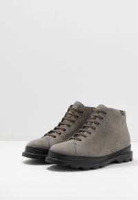 Bottes chelsea en suède gris avec semelle en caoutchouc noire, design à lacets, bout rond et détails de couture contrastants. Surface texturée et lignes épurées.