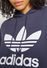 Granatowa bluza z kapturem z dużym białym logo trefoil Adidas i poziomymi paskami. Posiada kaptur z troczkami i miękką fakturę tkaniny.