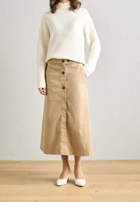 Beige bomulls midi kjol med knäppning fram, fickor på sidan och en lätt utsvängning i nederkanten, i kombination med en vit ribbad tröja och vita klackar.