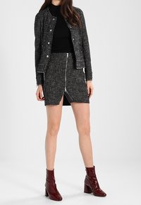 Veste en tweed noir et blanc avec boutons en argent, associée à un haut noir et une jupe en tweed noir zippée, complétée par des bottines bordeaux.