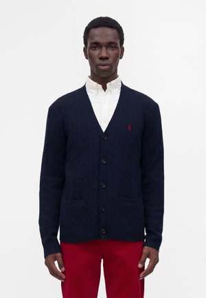 Homme portant un cardigan en maille torsadée bleu marine boutonné sur une chemise blanche et un pantalon rouge, debout devant un fond blanc uni.