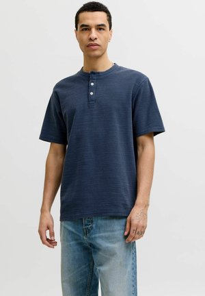 Mann trägt ein marineblaues Henley-Shirt mit kurzen Ärmeln und drei Knöpfen sowie hellblaue Jeans und steht vor einem schlichten Hintergrund.