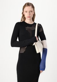 Svart stickad klänning med utskickade mönster, långa ärmar; en ärm har en blå accent. Vit väska som hålls med ett band.