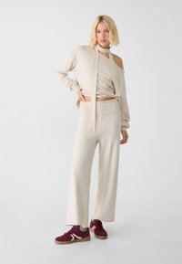 Top corto in maglia beige con scollatura asimmetrica e una lunga cintura, abbinato a pantaloni beige a gamba larga e sneakers in suede bordeaux con dettagli bianchi.