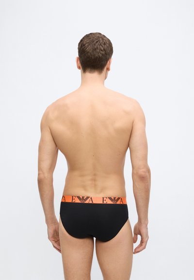 Άνδρας μοντέλο, φωτογραφημένος από πίσω, φορά μαύρα εσώρουχα με πορτοκαλί μέση που φέρει μαύρα λογότυπα και γράμματα, στέκεται μπροστά σε λευκό φόντο.