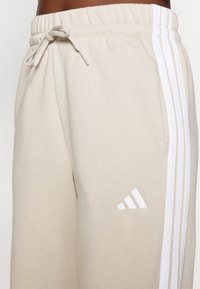 Beige sporta bikses ar auklu viduklī, ar trim baltām svītrām sānos un baltu logo apakšējā kājā. Mīksta auduma tekstūra.