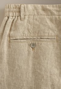 Pantalon en lin beige avec une taille élastique, une passants de ceinture et une poche passepoilée arrière sécurisée par un unique bouton.