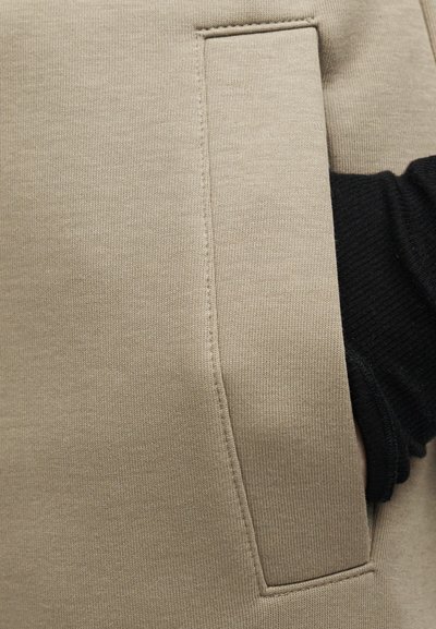 Χέρι με μαύρο μανίκι τοποθετημένο μέσα στην τσέπη μπεζ φούτερ με ορατή κάθετη ραφή και υφή υφάσματος.