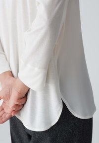 Blusa bianca leggera con orlo arrotondato e maniche a 3/4. Tessuto morbido, design minimale, abbinata a pantaloni grigio scuro.