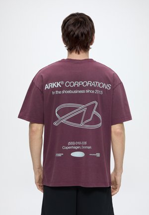 Osoba nosząca bordową koszulkę ARKK CORPORATIONS z białym napisem i logo, stojąca tyłem do kamery na białym tle.