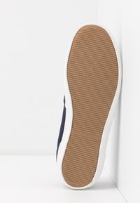 Basket décontractée unique avec semelle en caoutchouc marron texturée orientée vers l'avant, bords blancs et tige en tissu bleu foncé sur fond blanc d'angle.