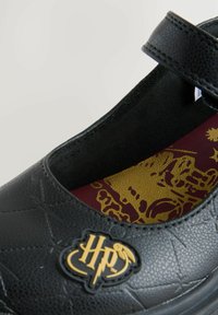 Chaussure Mary Jane noire en synthétique avec une bride, dotée d'un motif texturé, d'un logo doré "HP" et d'une semelle intérieure à motif rouge et or.