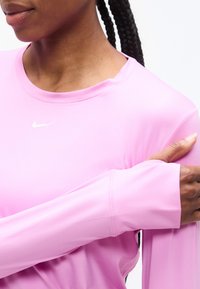 Άτομο με σκούρο δέρμα και πλεξούδες στα μαλλιά, που φοράει ροζ ανοιχτό μακρυμάνικη μπλούζα Nike, κρατώντας τον αριστερό βραχίονα με το δεξί χέρι.