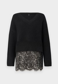 Pull en maille noire à col en V avec manches longues et un ourlet festonné en dentelle qui dépasse le bas du pull.