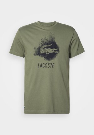 Olivově zelené bavlněné tričko se černou grafikou krokodýla a textem "LACOSTE" níže. Krátké rukávy a klasický kulatý výstřih.
