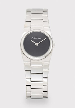 Montre-bracelet en métal argenté avec un cadran rond noir. Ses caractéristiques incluent deux aiguilles argentées et le texte "Calvin Klein" sur le cadran.