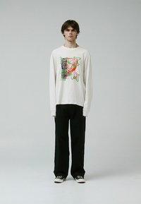 Camisa blanca de manga larga con diseño gráfico abstracto multicolor; combinada con pantalones negros holgados y zapatillas negras.